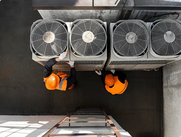 Top-down view of two workers in orange safety vests inspecting large rooftop industrial HVAC fan units