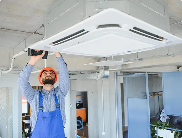 A technician in an orange hard hat using a power drill to install a ceiling cassette air conditioner in an office