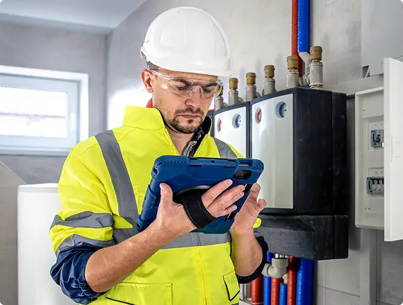 An engineer in a white hard hat and high-visibility jacket using a tablet to inspect a boiler room with pipes and valves