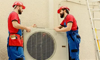 Two technicians in red uniforms and hard hats installing an outdoor air conditioning unit on a building wall
