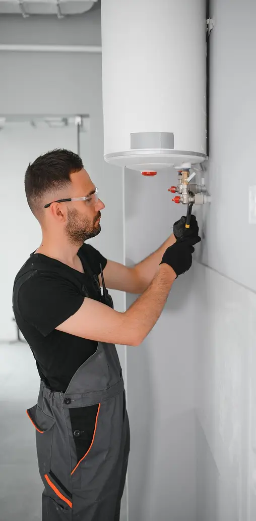 A plumber in overalls and safety glasses installing or servicing a wall-mounted electric water heater