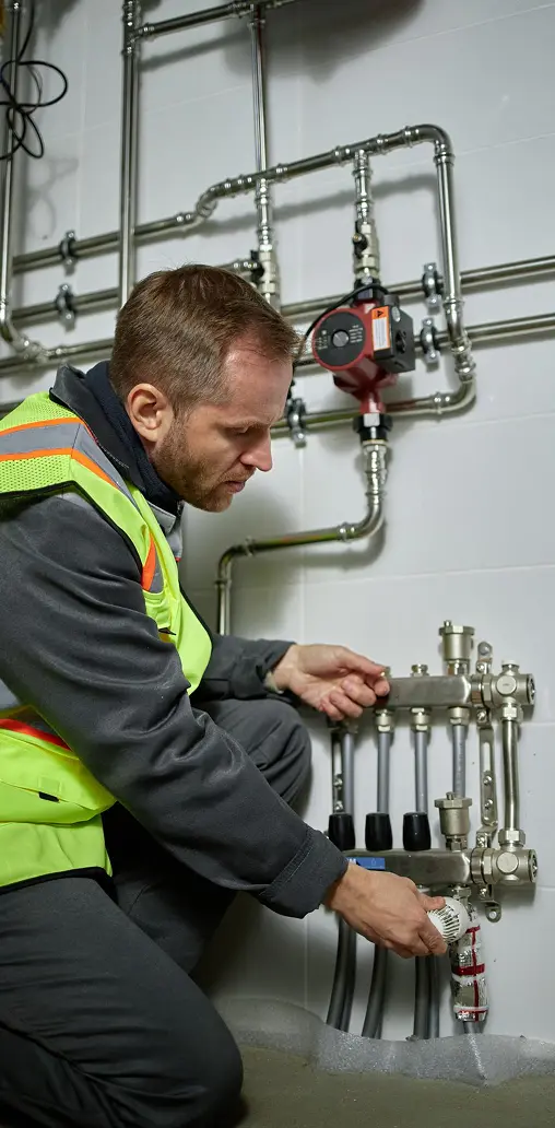 A technician in a high-visibility vest adjusting valves on a heating manifold system with metal pipes on a wall