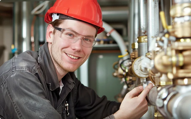 Smiling engineer in a red hard hat and safety goggles adjusting industrial pipe valves