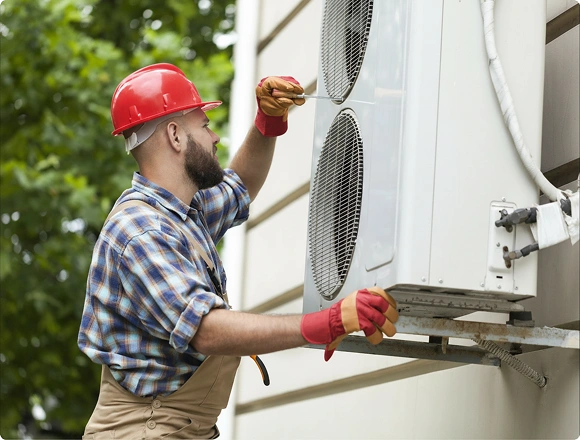 HVAC technician in a red hard hat installing an outdoor air conditioning unit on a building exterior