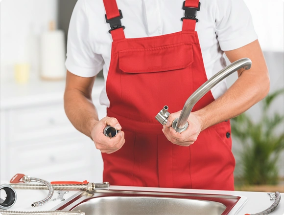 Plumber in red overalls holding a stainless steel faucet and connector while installing it on a kitchen sink, with braided supply hoses in the foreground