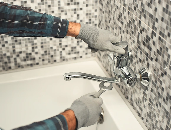 Gloved plumber using a wrench to install a chrome wall-mounted faucet on a bathtub against a mosaic tile wall