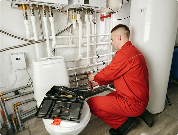 Plumber in red coveralls repairing pipes in a utility room with an open toolbox nearby