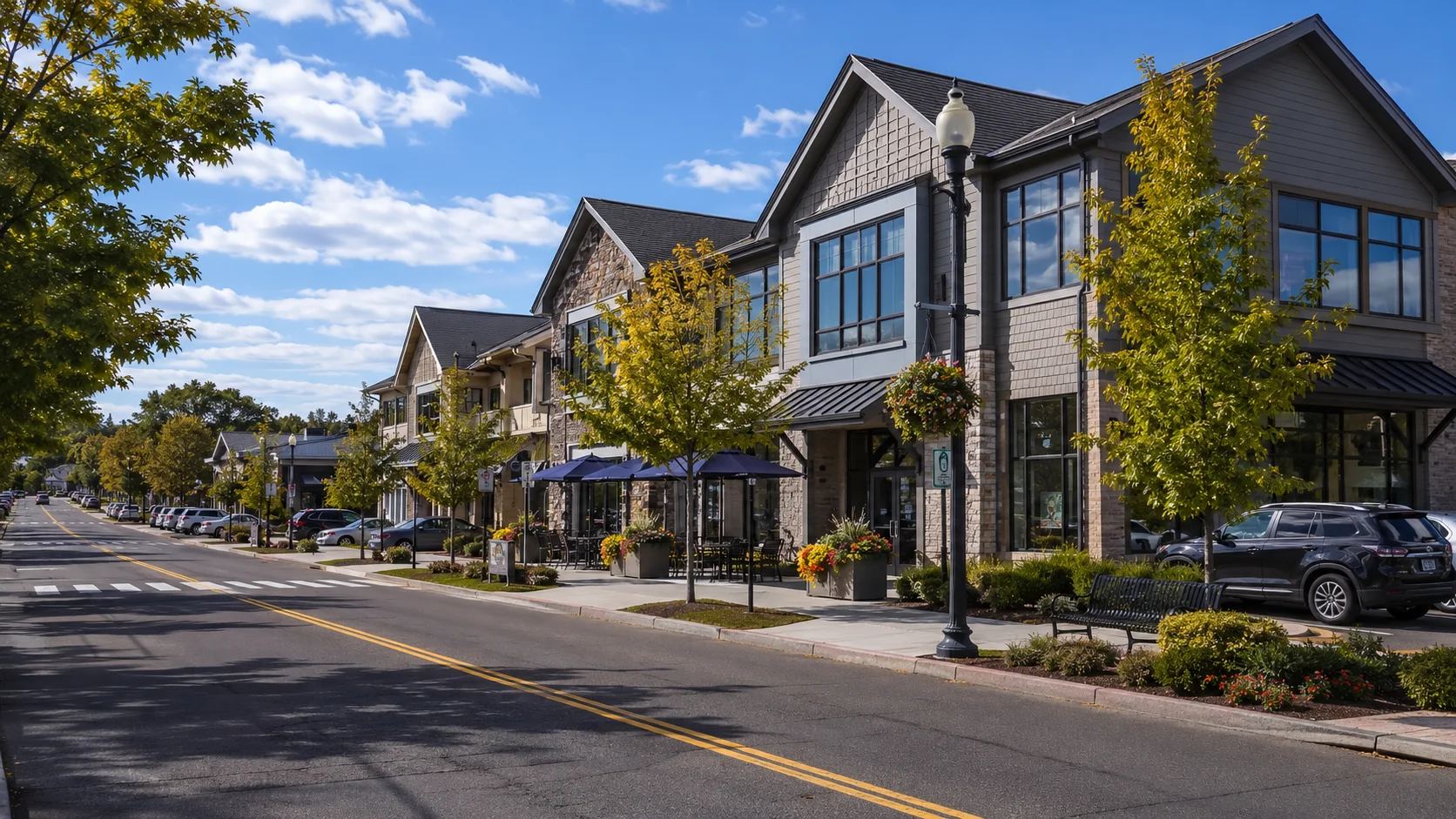 Sunny suburban shopping street with modern two-story buildings, young trees, and parked cars along an empty road.