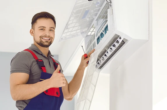 Smiling HVAC technician in blue overalls giving a thumbs-up while servicing a wall-mounted air-conditioning unit.