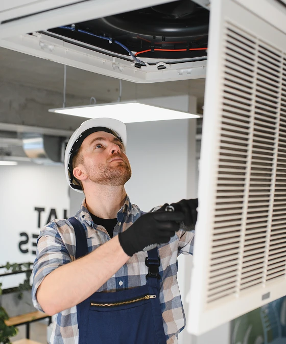 Male technician wearing a hard hat and work gloves examines an overhead HVAC vent while holding a wrench.